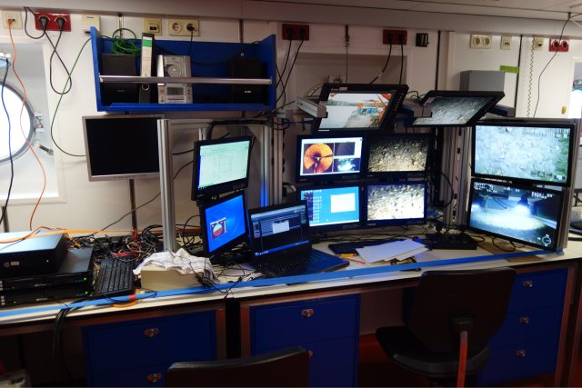 The ROV control centre, built in one of the workspaces below deck. The 80s design of the ship is clearly recognisable in the furnishings. Beige and white walls, only pierced by portholes, and furniture in the blue of the ship's hull exude an atmosphere that is simple but sufficient for working. Countless screens, computers, recording devices and control units are secured against high seas with a multitude of tension belts. The monitors show the images, navigation and orientation data of the cameras, the sonar and the positioning.