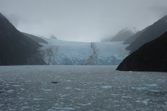 Gloomy weather at the end of Garibaldi Fjord, where the glacier extends from its valley led to the waterline, where it dissolves in a blanket of ice floes on the water surface. Dense clouds obscure the view of the mountain range in the background, so that only the nearby fjord flanks are visible. The little daylight that makes it through the hazy air allows hardly any colours to be discerned, which is why the blue-grey shades of the rock faces on both sides appear brighter and less contrasted with increasing distance.