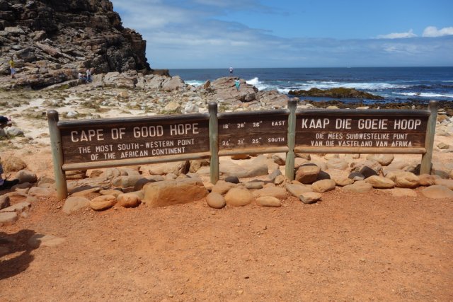 The southernmost tip of Africa. A view over the famous three-part wooden sign, which reads in large letters Cape of good hope; The most south-western point of the african continent in English and Afrikaans. In the middle are the geographical coordinates of this place: 18 degrees 28 minutes 26 seconds East and 34 degrees 21 minutes 25 seconds South. It is a warm, sunny day with little wind. The sea in the background is relatively calm and only a few whitecaps appear where the waves meet the rocky coastline that stretches up to the shield where it turns into rusty red sand.