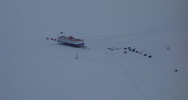 Aerial view of the German Antarctic station Neumayer III of the Alfred-Wegener-Institute at the edge of Atka Bay. The view from the aircraft on approach makes the station look like a miniature model. The view is obscured by a closed cloud cover, so that no horizon is visible. Nevertheless, the countless snowmobiles, snowscooters, outdoor shelters as well as the wind turbine and the antenna of the satellite connection line up as small dots around the station. Faintly but still as a slightly grey contrast, paths spread out from the station. The station itself, with its distinctive red-white-blue painting, is a rectangular block that seems to float above the ice surface, supported by pillars.