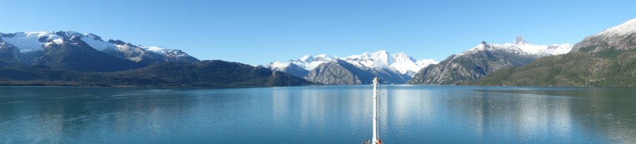 Panoramic view of the fjord landscape of Tierra del Fuego. A sunny, cloudless and almost windless day. The snow-covered mountain peaks shine in the sunlight in a multi-faceted way, first changing into rocky grey until they finally end in green vegetation that reaches down to the water. Only the front tip of the mast is visible from the ship, indicating the course over the flat surface of the water, in which the landscape and the blue sky are blurred.