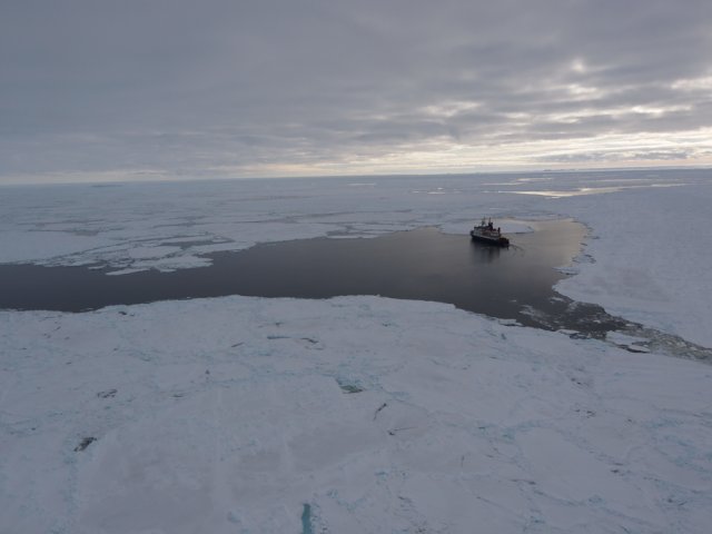 The bird's eye view shows the Polarstern in a small ployna of water otherwise covered by sea ice. The sky is covered with thick clouds and only a narrow strip on the horizon lets a little daylight into the scenery.