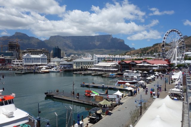 View over the Waterfront, Cape Town's harbour promenade, to the city's landmark, the imposing Table Mountain in the background. Over the plateau of the mountain, which looks as if it has been cut off with a knife, white fluffy clouds pass through the otherwise azure summer sky. The foreground is dominated by the harbour basin, where only a few boats are moored. The pier is moderately busy, with only a few visitors strolling along the promenade, on whose waterfront various stalls offer their seafood and a wide variety of souvenir items. At the corner of the basin, the Ferris wheel, which makes the Waterfront so distinctive, towers up and offers a magnificent view of the city's coastline. From there on, restaurants line the waterfront, eventually being replaced by historic buildings.