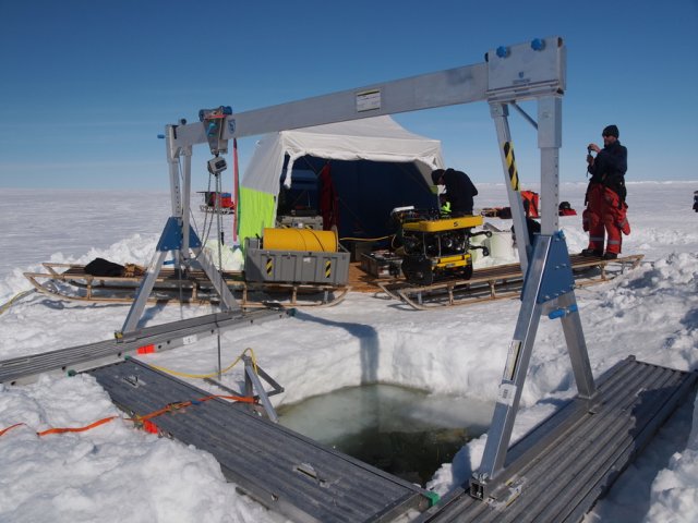 ROV station on the sea ice of Drescher Inlet. An aluminium sliding beam is set up above the ice hole, which measures about two and a half square metres. To prevent its side parts from sinking into the snow, they are placed on planks. Behind the square hole stands the ROV and the associated winch on two Nansen sleds. The ROV itself measures about sixty by sixty by eighty centimetres and belongs to the observation class of underwater robots. This means that it is not designed for heavy work and is mainly used for monitoring. The yellow buoyancy bodies are arranged at the top and bottom, enclosing the entire technology. The winch standing next to it accommodates a six hundred metre long bright yellow cable and weighs over a hundred and twenty kilos alone. A white and yellow tent is erected in the background, housing the control centre. As on so many days, the sky is cloudless and the sun, high in the zenith, casts only short shadows on the scenery.