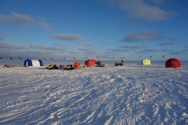 The finished base at Drescher Inlet. The sun is now low over the horizon, casting long shadows in a warm light. Small fleecy clouds line the sky. The camp consists of tents and igloos. From left to right comes first the ROV workshop and my accommodation, a blue and white semi-tubular tent with a proper entrance door. Next comes our opportunity for private needs, a pyramid-shaped Scott tent in bright red. The communal igloo is in the middle of the base. To the right is the generator bank, a white and yellow rectangular tent and another red igloo. The yellow snowmobiles with their Nansen sleds attached are parked in front of the camp.