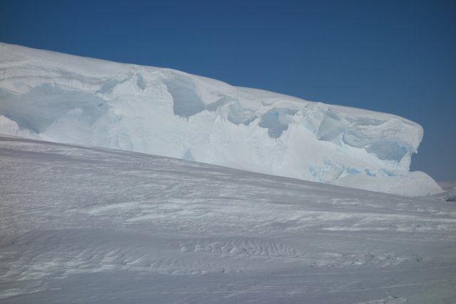 Photo of the ice shelf wall from the perspective of the inlet, which stretches up to twenty metres into the sky. The wind has brought out artistic sculptures so that thick waves of white snow spill over the top of the ice wall like mountains of whipped cream on a piece of cake.