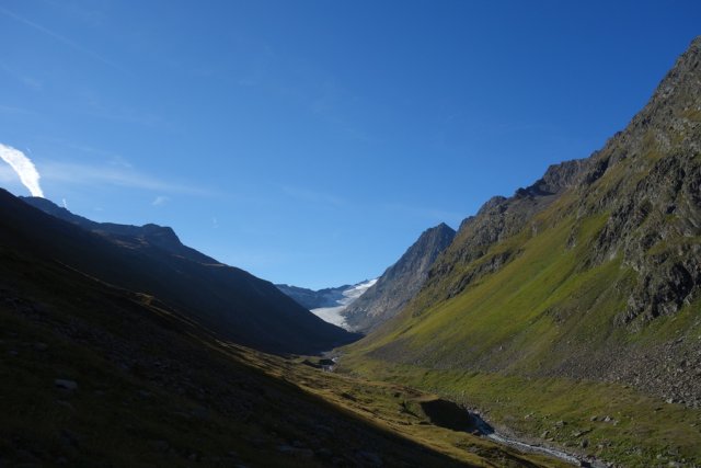 View up to the Gurgler Ferner above the vegetation border. The parabolic valley cross-section was formed by the once massive glacier, which has retreated to the highest regions of the mountains over the millennia. In the foreground, the slopes are still covered with shimmering green grasses, leaving the stage to the greyish rock and lighter ice higher up. The morning sun shines flatly, illuminating only the right flank of the valley, while the left side lies entirely in shadow.