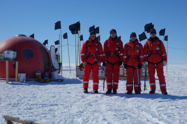 Our team shortly before the first ice exploration. Once again, a cloudless azure sky. Despite the warming sun, we are dressed in our red thermal suits, wearing caps and snow goggles to protect us from the merciless UV rays. We are standing in front of our kitchen igloo, a red, spherical GRP dwelling, also called a red tomato because of its colour and shape. Bamboo poles with black flags mark everything that could be covered by snow drift.